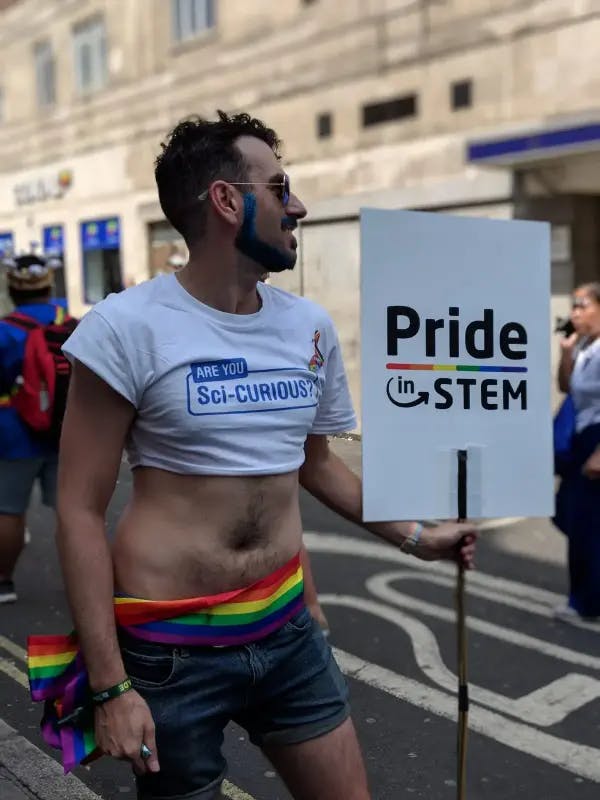 Alfredo wearing a crop-top, rainbow flag round his waist and short shorts holding a placard that reads "Pride in STEM"